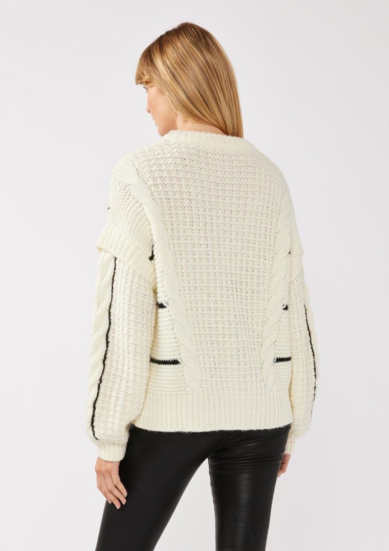 [Color: Cream/Black] A back facing image of a blonde model wearing a cozy knit sweater with a mixed fabric patchwork and cable knit detailing. This casual sweater features a crew neckline, long sleeves with a dropped shoulder, and a contrasting stripe accent. A cold weather staple for the fall and winter season. 