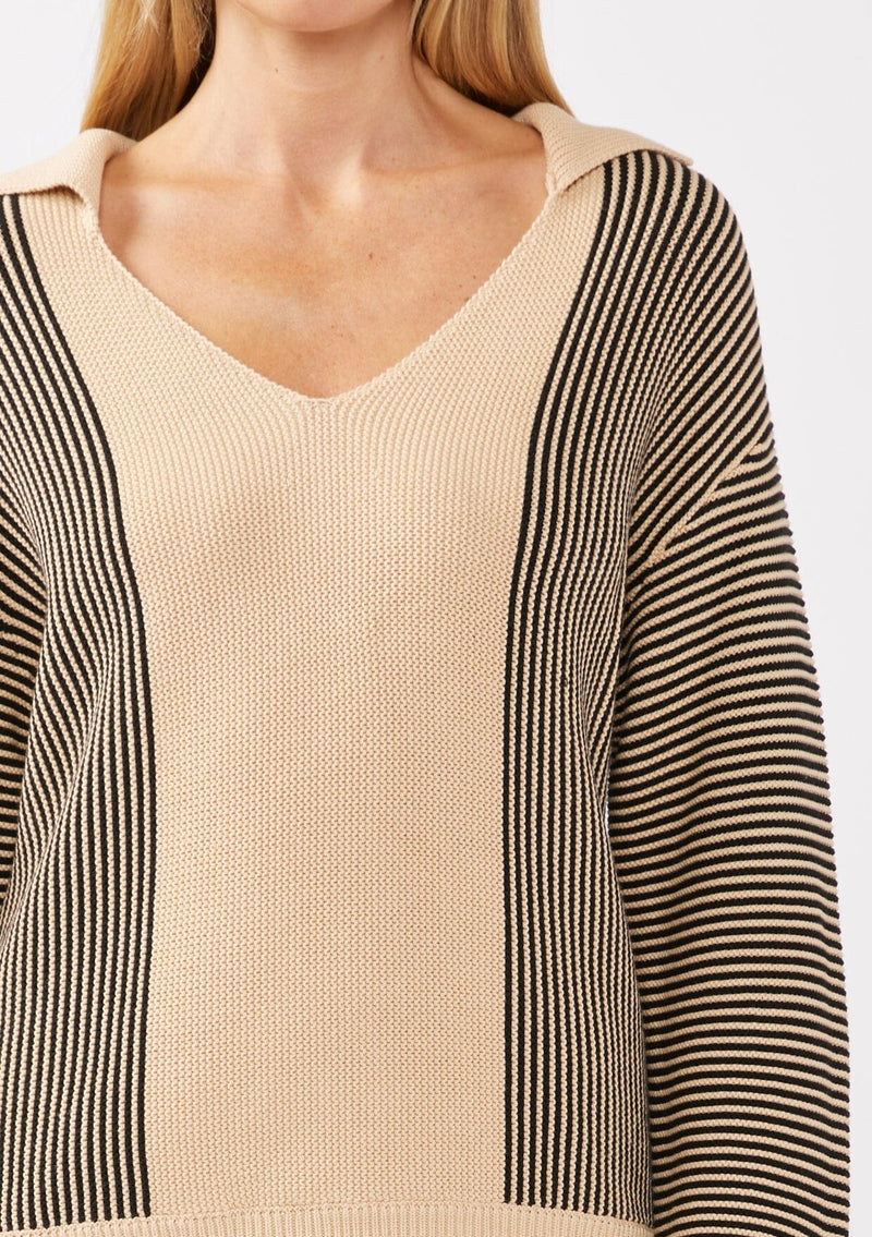 [Color: Natural/Black] A detail image of a blonde model wearing a striped knit pullover with a bold contrast. Featuring a collared v-neckline, long sleeves, dropped shoulders, and a ribbed cuffs and hem. A relaxed knit sweater styled with black bottoms for the fall season. 