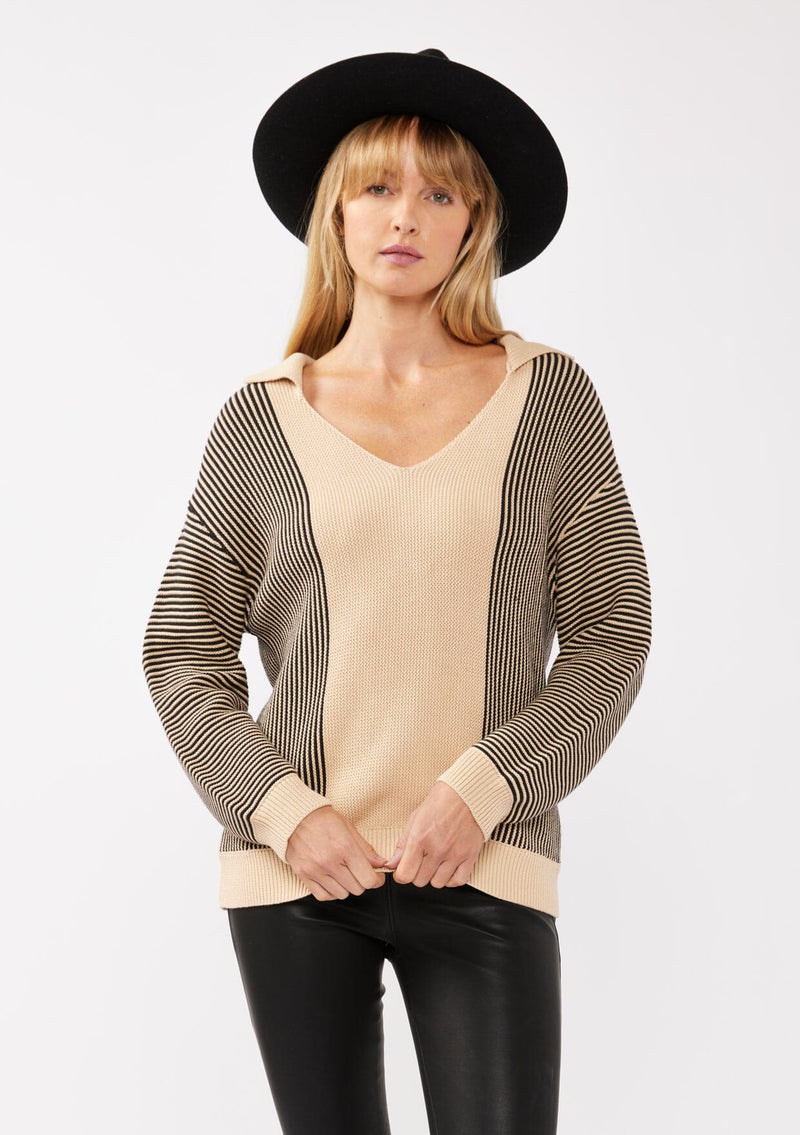 [Color: Natural/Black] A front facing image of a blonde model wearing a striped knit pullover with a bold contrast. Featuring a collared v-neckline, long sleeves, dropped shoulders, and a ribbed cuffs and hem. A relaxed knit sweater styled with black bottoms for the fall season. 