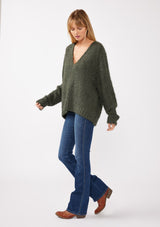 [Color: Olive] A side-facing image of a blonde model wearing a green cozy, fuzzy popcorn-textured pullover sweater with long sleeves, a classic V-neckline, and a high-low hemline. Ideal for lounging or casual fall outings.