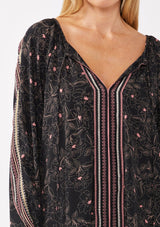 [Color: Black/Rose] A model wearing a classic black bohemian peasant top with romantic pink floral print. With long voluminous sleeves and a split v neckline with tassel ties. 