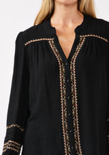 [Color: Black/Mocha] A detail image of a blonde model wearing a black button-front blouse with contrasting embroidered details throughout. The boho blouse features a relaxed fit, long sleeves, a split V-neckline, and button-cuff closures. Styled with denim for the perfect fall ensemble. 