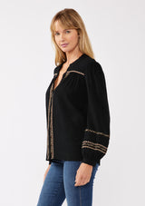 [Color: Black/Mocha] A side facing image of a blonde model wearing a black button-front blouse with contrasting embroidered details throughout. The boho blouse features a relaxed fit, long sleeves, a split V-neckline, and button-cuff closures. Styled with denim for the perfect fall ensemble. 