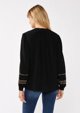 [Color: Black/Mocha] A back facing image of a blonde model wearing a black button-front blouse with contrasting embroidered details throughout. The boho blouse features a relaxed fit, long sleeves, a split V-neckline, and button-cuff closures. Styled with denim for the perfect fall ensemble. 