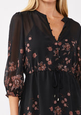 [Color: Black/Dusty Rose] A detail image of a blonde model wearing a black chiffon mini dress with a pink floral print. The boho-inspired design features a split V-neckline, button front, 3/4 sleeves, and an adjustable drawstring waist. Paired with long suede boots for a casual yet polished fall look.