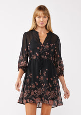 [Color: Black/Dusty Rose] A front faccing image of a blonde model wearing a black chiffon mini dress with a pink floral print. The boho-inspired design features a split V-neckline, button front, 3/4 sleeves, and an adjustable drawstring waist. Paired with long suede boots for a casual yet polished fall look.