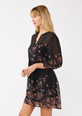 [Color: Black/Dusty Rose] A side image of a blonde model wearing a black chiffon mini dress with a pink floral print. The boho-inspired design features a split V-neckline, button front, 3/4 sleeves, and an adjustable drawstring waist. Paired with long suede boots for a casual yet polished fall look.