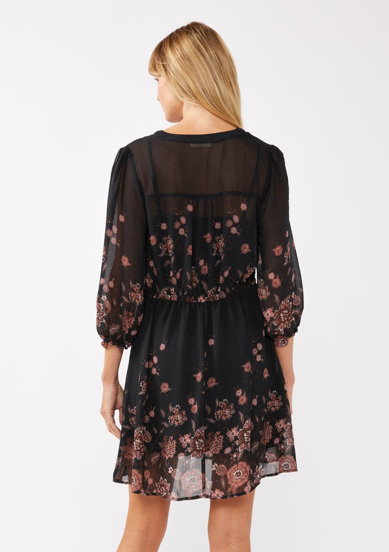 [Color: Black/Dusty Rose] A back facing image of a blonde model wearing a black chiffon mini dress with a pink floral print. The boho-inspired design features a split V-neckline, button front, 3/4 sleeves, and an adjustable drawstring waist. Paired with long suede boots for a casual yet polished fall look.