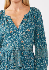 [Color: Teal/Cream] A detail image of a blonde model wearing an all-over teal floral print midi dress. The dress features long sleeves, a button-front, a self-tie waist belt, and a straight hem. Styled with brown boots, it highlights a relaxed silhouette and effortless elegance.