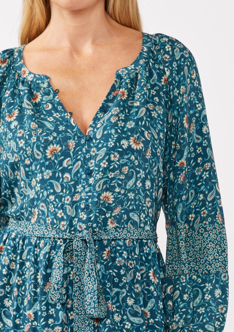 [Color: Teal/Cream] A detail image of a blonde model wearing an all-over teal floral print midi dress. The dress features long sleeves, a button-front, a self-tie waist belt, and a straight hem. Styled with brown boots, it highlights a relaxed silhouette and effortless elegance.