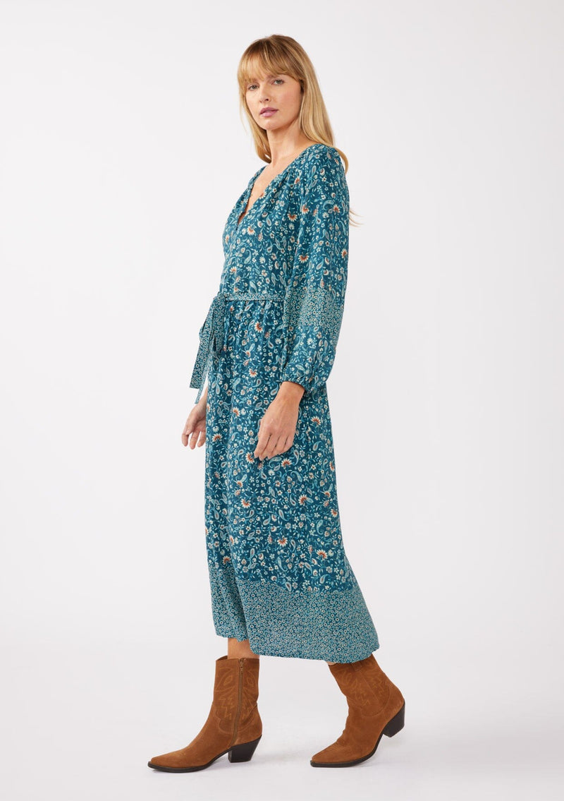 [Color: Teal/Cream] A full body side image of a blonde model wearing an all-over teal floral print midi dress. The dress features long sleeves, a button-front, a self-tie waist belt, and a straight hem. Styled with brown boots, it highlights a relaxed silhouette and effortless elegance.