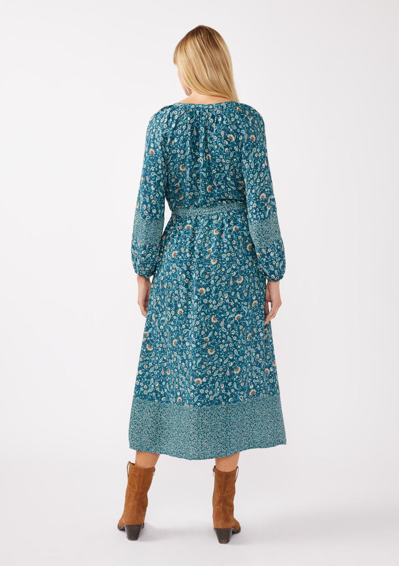 [Color: Teal/Cream] A back facing image of a blonde model wearing an all-over teal floral print midi dress. The dress features long sleeves, a button-front, a self-tie waist belt, and a straight hem. Styled with brown boots, it highlights a relaxed silhouette and effortless elegance.