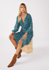 [Color: Teal/Cream] A sitting image of a blonde model wearing an all-over teal floral print midi dress. The dress features long sleeves, a button-front, a self-tie waist belt, and a straight hem. Styled with brown boots, it highlights a relaxed silhouette and effortless elegance.