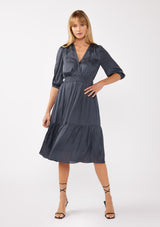 [Color: Midnight] A front facing image of a blonde model wearing a grey blue satin midi dress with a surplice v-neckline, 3/4 sleeves, and  a flowy tiered skirt. Complete with a stretchy smocked waistline and a button snap closure at the neckline. Perfect for any holiday event or special occasion. 