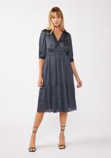 [Color: Midnight] A front facing image of a blonde model wearing a grey blue satin midi dress with a surplice v-neckline, 3/4 sleeves, and  a flowy tiered skirt. Complete with a stretchy smocked waistline and a button snap closure at the neckline. Perfect for any holiday event or special occasion. 