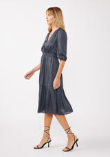 [Color: Midnight] A side facing image of a blonde model wearing a grey blue satin midi dress with a surplice v-neckline, 3/4 sleeves, and  a flowy tiered skirt. Complete with a stretchy smocked waistline and a button snap closure at the neckline. Perfect for any holiday event or special occasion. 