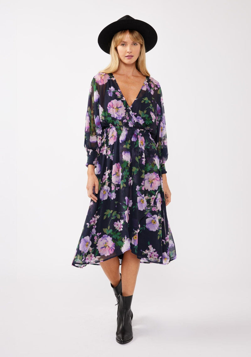 [Color: Navy/Lilac] A blonde model wearing a purple floral midi dress crafted in flowy chiffon. This faux wrap dress features a surplice v-neckline, flowy high low hemline, voluminous long sleeves, and a smocked waist and cuff. Perfect for special occasions and weddings. 