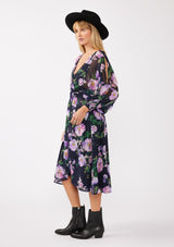 [Color: Navy/Lilac] A blonde model wearing a purple floral midi dress crafted in flowy chiffon. This faux wrap dress features a surplice v-neckline, flowy high low hemline, voluminous long sleeves, split sleeve detail, and a smocked waist and cuff. Perfect for special occasions and weddings. 