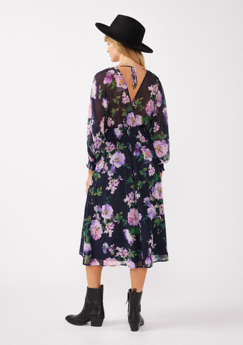 [Color: Navy/Lilac] A back facing image of a blonde model wearing a purple floral midi dress crafted in flowy chiffon. This faux wrap dress features a surplice v-neckline, flowy high low hemline, voluminous long sleeves, split sleeve detail, and a smocked waist and cuff. Perfect for special occasions and weddings. 