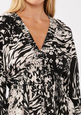 [Color: Black/Cream] A detail image of a blonde model wearing a versatile black and ivory multi print mini dress. This long sleeve boho mini dress features a smocked neckline, smocked empire waist, and side pockets. Perfect for casual outings, vacation, or dinner dates.  