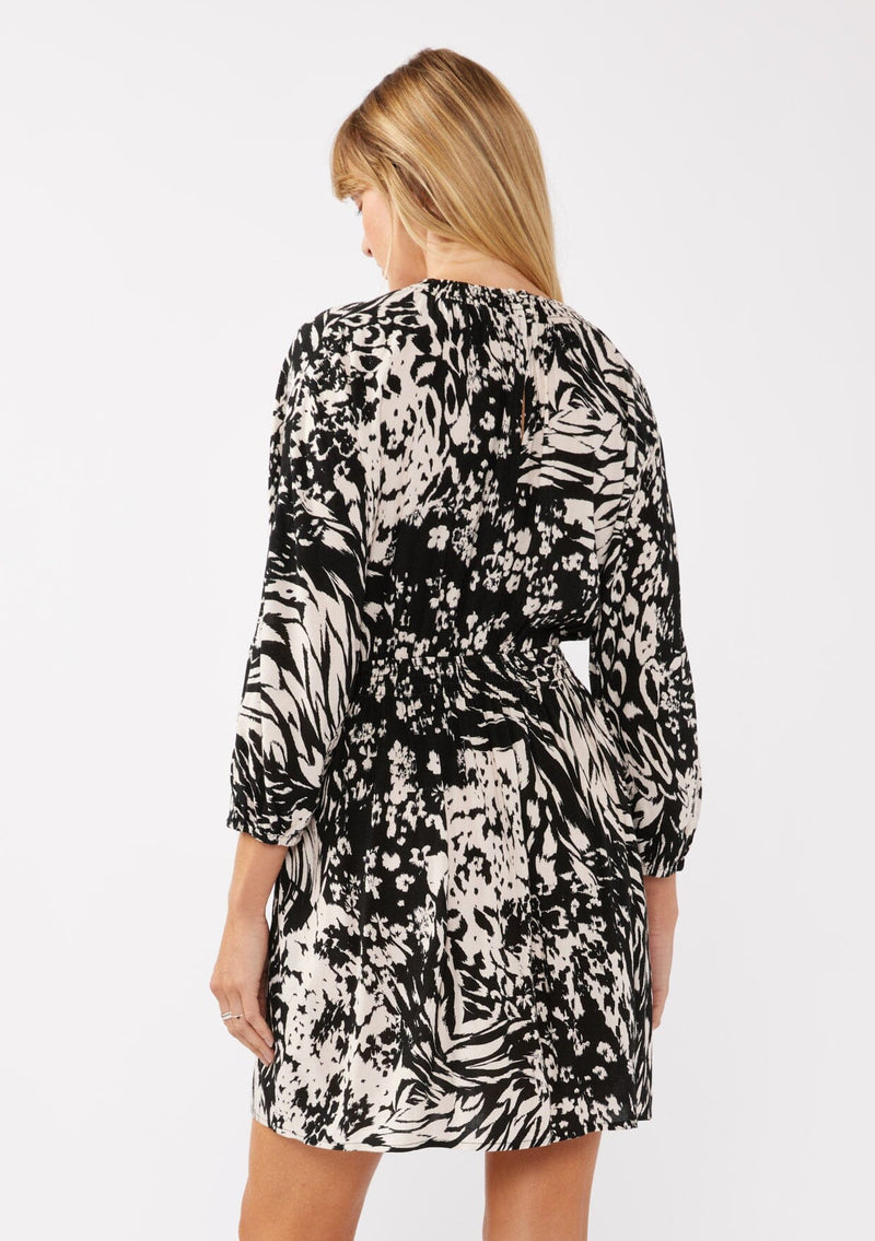 [Color: Black/Cream] A back facing image of a blonde model wearing a versatile black and ivory multi print mini dress. This long sleeve boho mini dress features a smocked neckline, smocked empire waist, and side pockets. Perfect for casual outings, vacation, or dinner dates.  