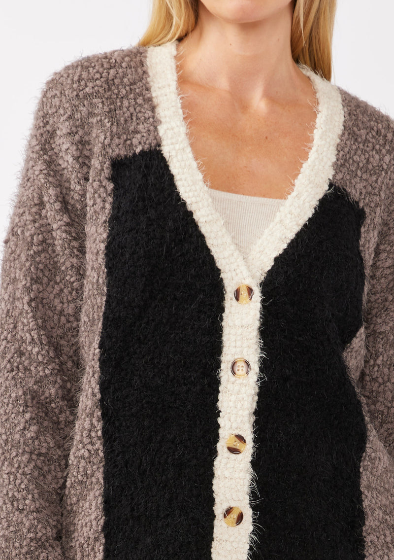 [Color: Vanilla Black] A detail image of a blonde model wearing an ultra warm color block cardigan in a soft fuzzy popcorn texture. Featuring a classic tortoiseshell button up front, a flattering v neckline, and a contrast trim. This fuzzy cardigan is perfect for layering during the fall season. 