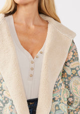 [Color: Midnight/Blush] A detail image of a blonde model wearing a green paisley and floral print jacket with a sherpa lining. A boho statement jacket with long sleeves, patch pockets, a hoodie, and a cozy draped silhouette for warmth and comfort. Styled with denim for a casual fall or winter look. 