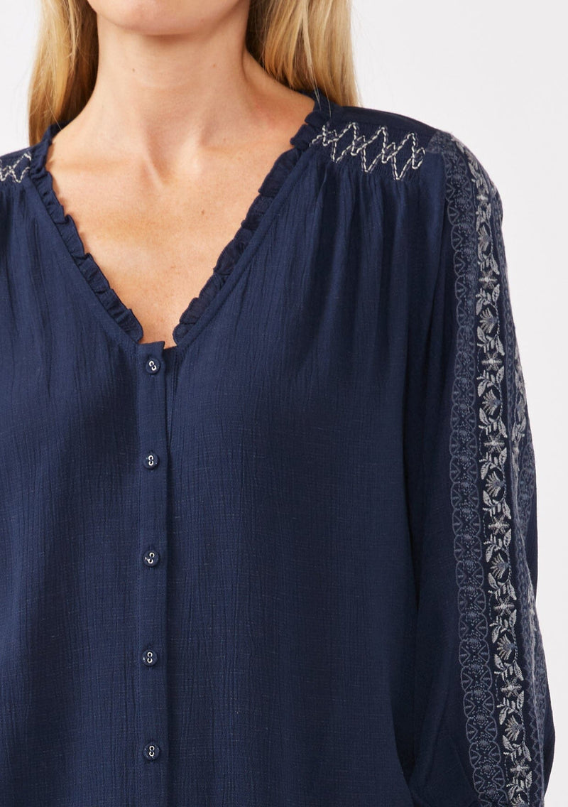 [Color: Navy] A detail image of a blonde model wearing an embroidered, navy blue blouse with a button front, ruffle trimmed v-neckline, 3/4 sleeves, and elastic wrist cuffs. Paired with brown corduroy bottoms for a laid back fall look. 