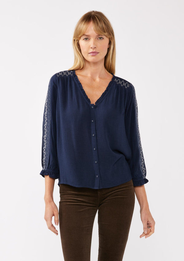 [Color: Navy] A front facing image of a blonde model wearing an embroidered, navy blue blouse with a button front, ruffle trimmed v-neckline, 3/4 sleeves, and elastic wrist cuffs. Paired with brown corduroy bottoms for a laid back fall look. 