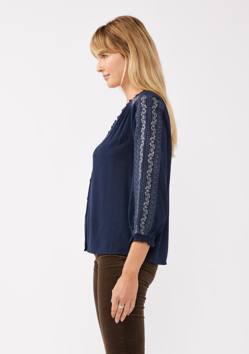 [Color: Navy] A side facing image of a blonde model wearing an embroidered, navy blue blouse with a button front, ruffle trimmed v-neckline, 3/4 sleeves, and elastic wrist cuffs. Paired with brown corduroy bottoms for a laid back fall look. 