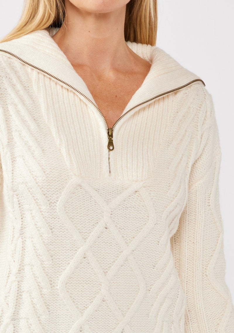 [Color: Natural] A detail image of a blonde model wearing a cozy cable knit pullover sweater in off white. This casual fall knit features a front half zipper front, long sleeves, and a rib trim hem and cuffs. 