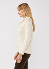 [Color: Natural] A side facing image of a blonde model wearing a cozy cable knit pullover sweater in off white. This casual fall knit features a front half zipper front, long sleeves, and a rib trim hem and cuffs. 