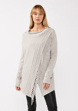 [Color: Heather Grey] A front-facing image of a blonde model wearing a grey fringed cardigan with an asymmetrical wrap design. The cardigan features hidden shoulder buttons for a wrap-front style, long sleeves, and intricate cable knit detailing.