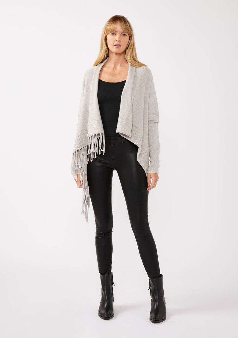 [Color: Heather Grey] A front-facing image of a blonde model wearing a grey fringed cardigan with an asymmetrical design. The cardigan features hidden shoulder buttons for a wrap-front style, long sleeves, and intricate cable knit detailing.