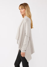 [Color: Heather Grey] A side-facing image of a blonde model wearing a grey fringed cardigan with an asymmetrical wrap design. The cardigan features hidden shoulder buttons for a wrap-front style, long sleeves, and intricate cable knit detailing.