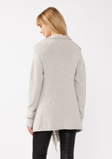 [Color: Heather Grey] A back-facing image of a blonde model wearing a grey fringed cardigan with an asymmetrical wrap design. The cardigan features hidden shoulder buttons for a wrap-front style, long sleeves, and intricate cable knit detailing.