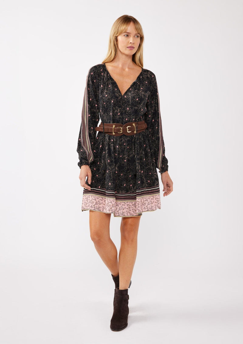 [Color: Black/Rose] A model wearing a classic black bohemian mini dress in a pink floral border print. With a loose, relaxed silhouette, long sleeves, and a split v neckline with tassels.