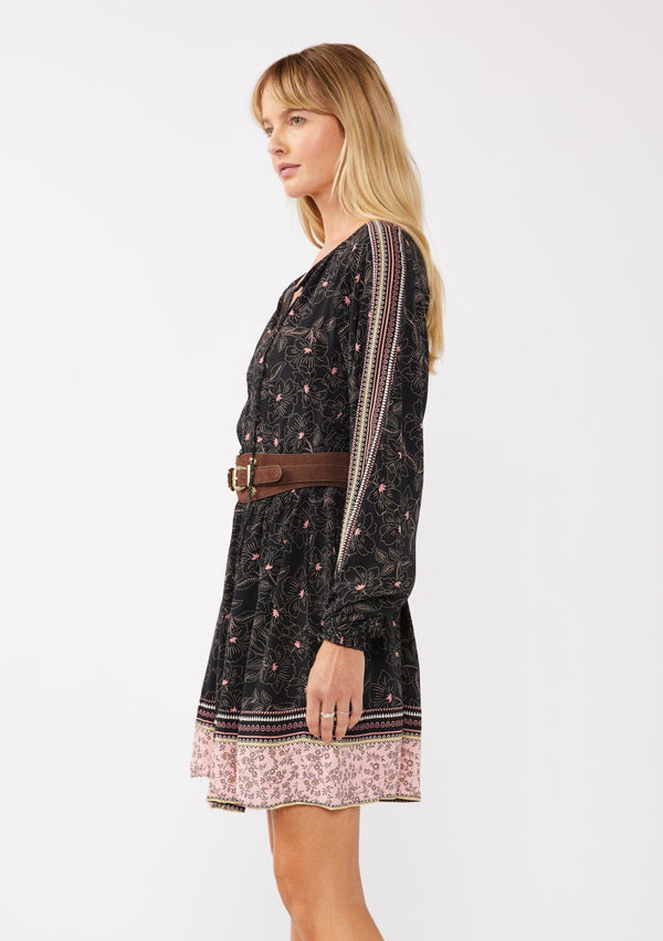 [Color: Black/Rose] A model wearing a classic black bohemian mini dress in a pink floral border print. With a loose, relaxed silhouette, long sleeves, and a split v neckline with tassels.