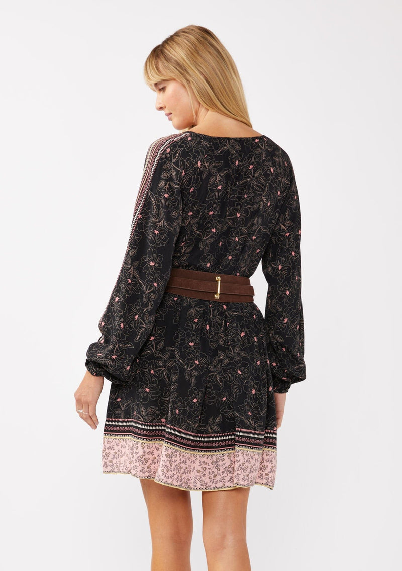 [Color: Black/Rose] A model wearing a classic black bohemian mini dress in a pink floral border print. With a loose, relaxed silhouette, long sleeves, and a split v neckline with tassels.