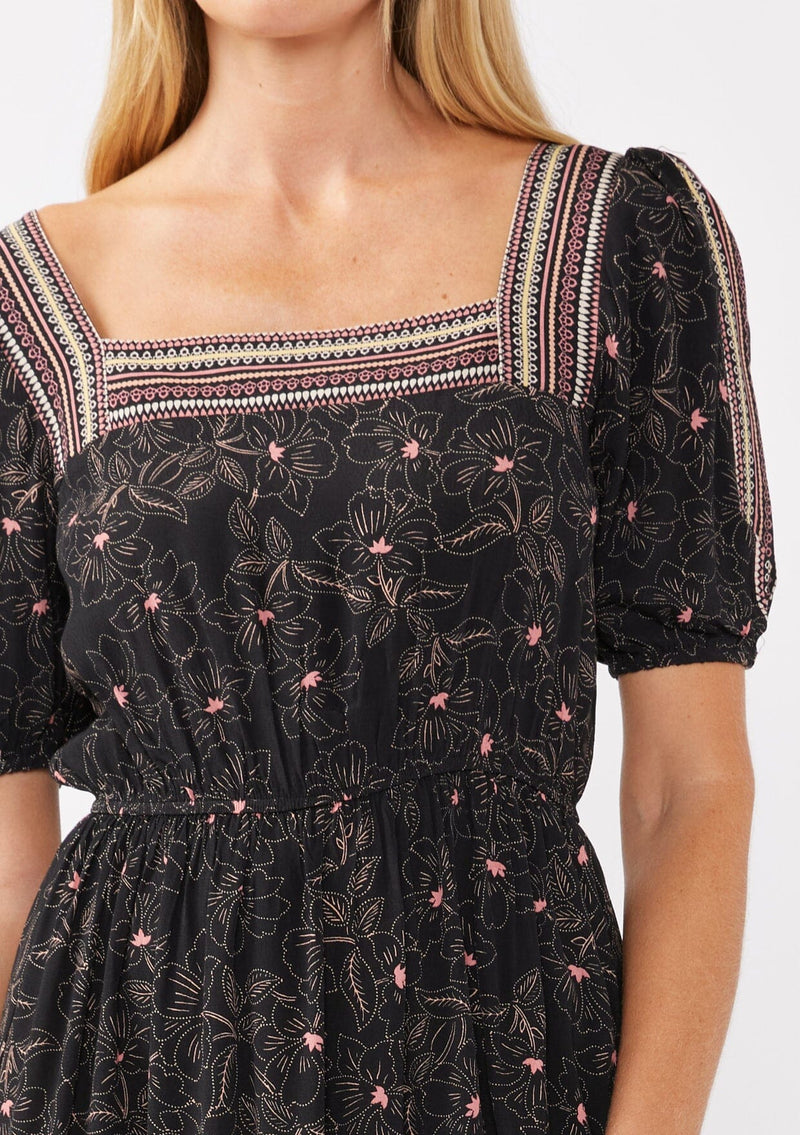 [Color: Black/Rose] A detail image of a blonde model wearing a floral bohemian maxi dress with a border print design. This flowy maxi features a square neckline, short puff sleeves,  and an elastic waist. 