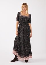 [Color: Black/Rose] A front facing image of a blonde model wearing a floral bohemian maxi dress with a border print design. This flowy maxi features a square neckline, short puff sleeves,  and an elastic waist. 