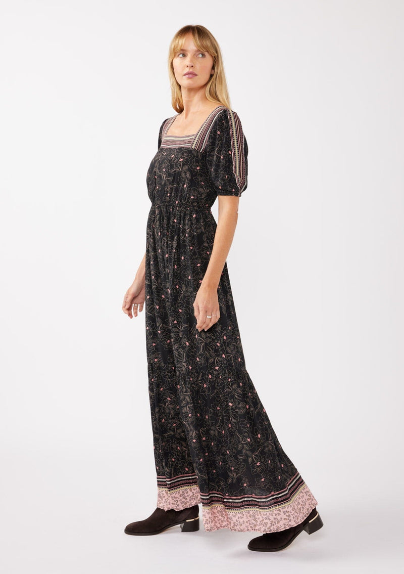 [Color: Black/Rose] A side facing image of a blonde model wearing a floral bohemian maxi dress with a border print design. This flowy maxi features a square neckline, short puff sleeves,  and an elastic waist. 