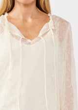 [Color: Ivory] A detail image of a blonde model wearing a crinkled sheer chiffon blouse with a floral pattern in ivory off white . This relaxed fit blouse features a double tie neck detail, long sleeves with ruffled elastic cuffs, and a detachable cami. 