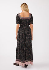 [Color: Black/Rose] A back facing image of a blonde model wearing a floral bohemian maxi dress with a border print design. This flowy maxi features a square neckline, short puff sleeves,  and an elastic waist. 