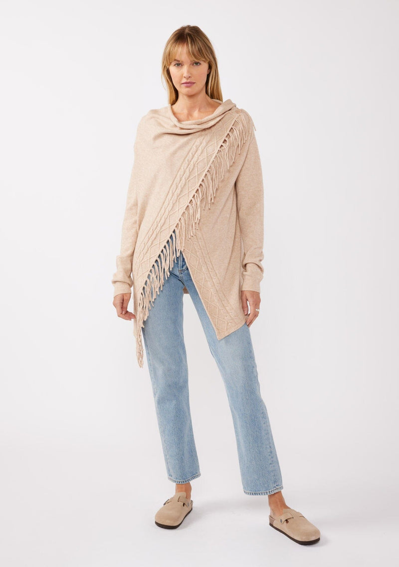 [Color: Heather Natural] A front-facing image of a blonde model wearing a brown tan fringed cardigan with an asymmetrical wrap design. The cardigan features hidden shoulder buttons for a wrap-front style, long sleeves, and intricate cable knit detailing.