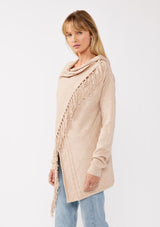 [Color: Heather Natural] A side facing image of a blonde model wearing a brown tan fringed cardigan with an asymmetrical wrap design. The cardigan features hidden shoulder buttons for a wrap-front style, long sleeves, and intricate cable knit detailing.