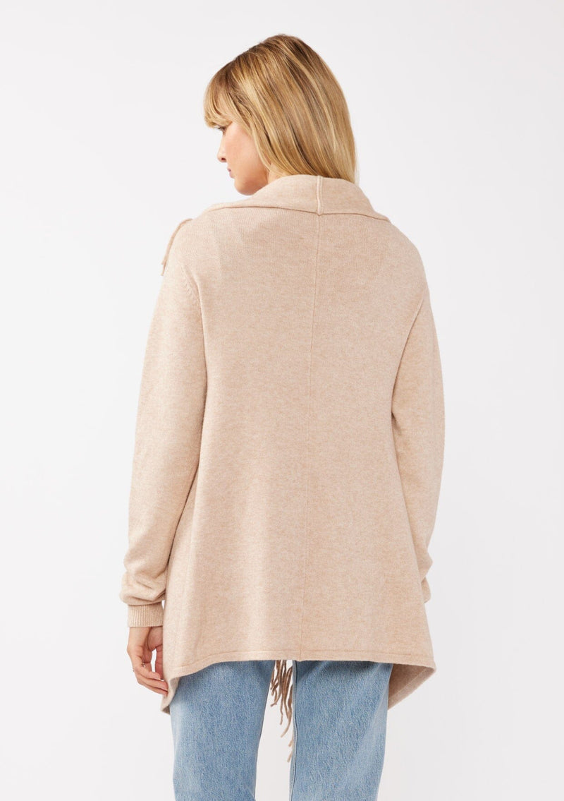 [Color: Heather Natural] A back facing image of a blonde model wearing a brown tan fringed cardigan with an asymmetrical wrap design. The cardigan features hidden shoulder buttons for a wrap-front style, long sleeves, and intricate cable knit detailing.