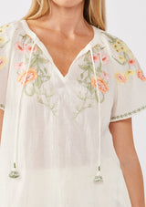 [Color: Cream/Coral] A detail image of a blonde model wearing a cotton cream top with contrasting floral embroidered details. This relaxed cotton top features short sleeves and a split v-neckline with ties. Perfect for lounging or vacations. 