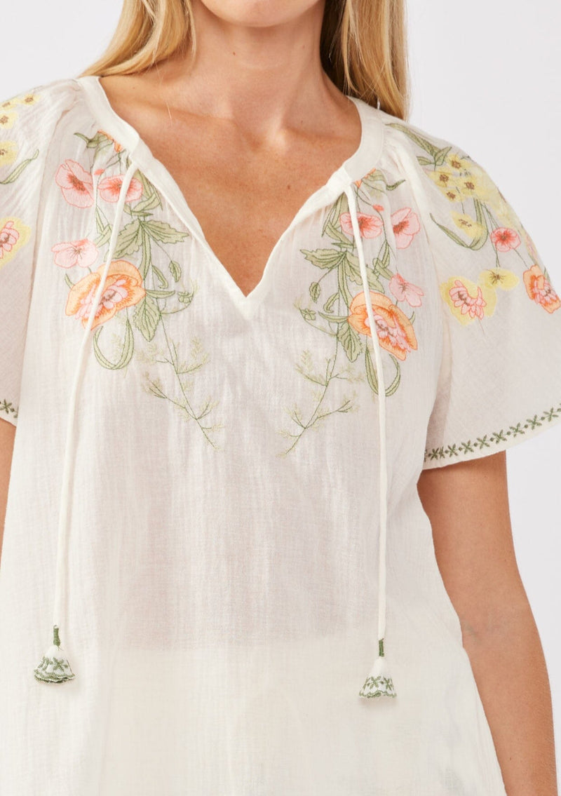 [Color: Cream/Coral] A detail image of a blonde model wearing a cotton cream top with contrasting floral embroidered details. This relaxed cotton top features short sleeves and a split v-neckline with ties. Perfect for lounging or vacations. 