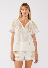 [Color: Cream/Coral] A front facing image of a blonde model wearing a cotton cream top with contrasting floral embroidered details. This relaxed cotton top features short sleeves and a split v-neckline with ties. Perfect for lounging or vacations. 